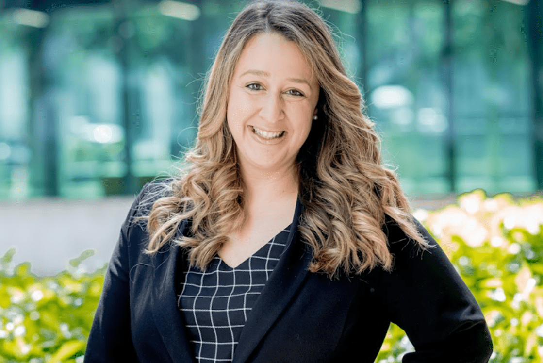 Image: Silvia is standing in front of a green bush. She is wearing a black blazer and black and white shirt. She has long blonde curly hair and is smiling at the camera.