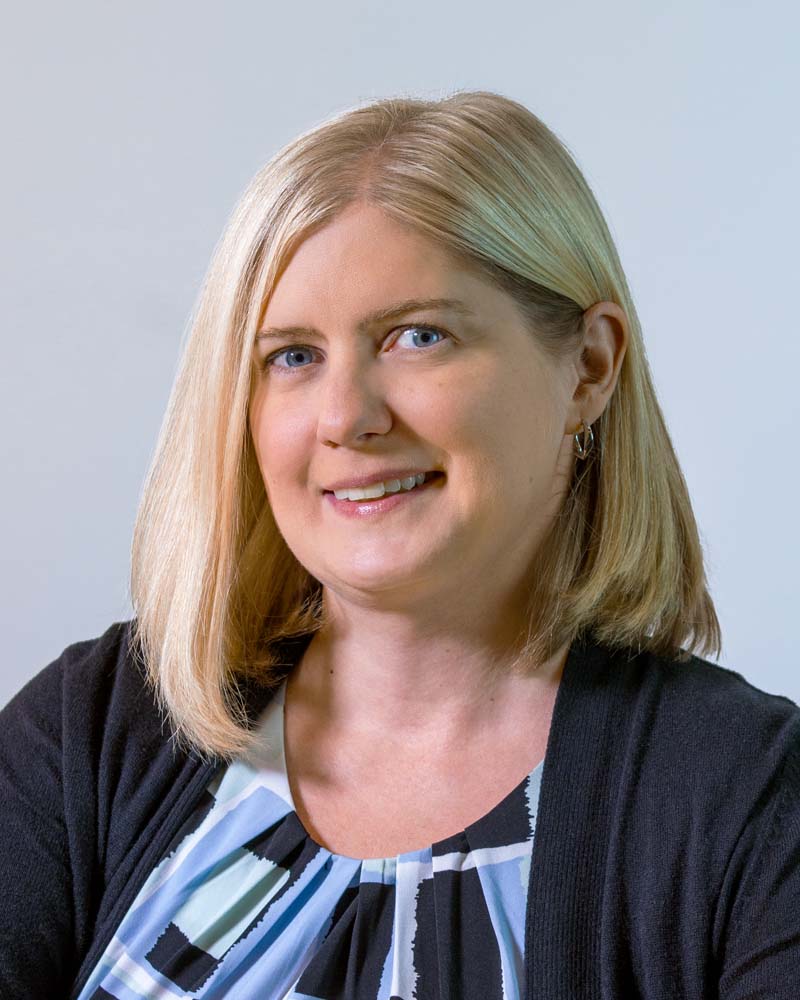 Image: Circular photo of Angela Dirk, a white woman with shoulder-length blonde hair and blue eyes, wearing a red and blue patterned shirt with a blue cardigan.