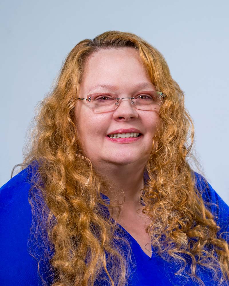 Image: Photo of Tanya Morrow, a white woman with light brown curly hair, pink lipstick, glasses, and a blue shirt, smiling.