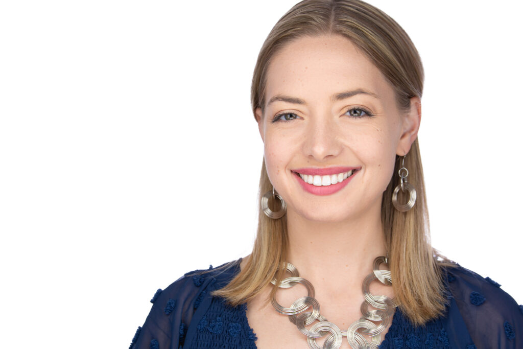 A woman in her 30's with light skin and shoulder-length blonde hair smiles at the camera. She wears a navy top and silver circular jewelry against a plain white background.