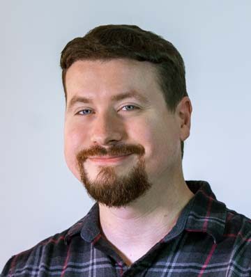 Image: Professional photo of Tyler Talcott, a white man with short brown hair, a goatee, wearing a plaid button down shirt.