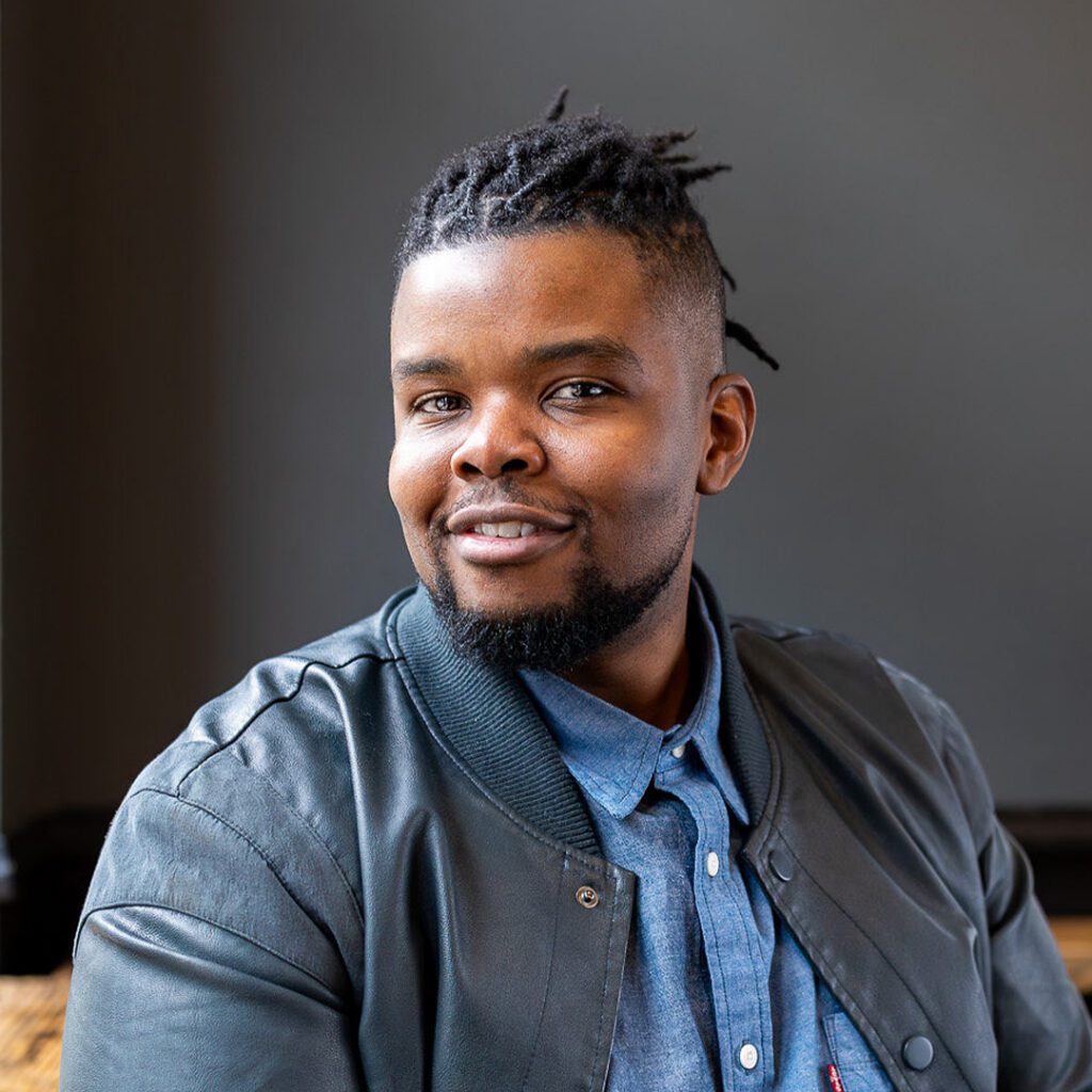 Photo of the presenter Paulo Bandua, a black male with locks smiling in a professional setting, wearing business casual attire.
