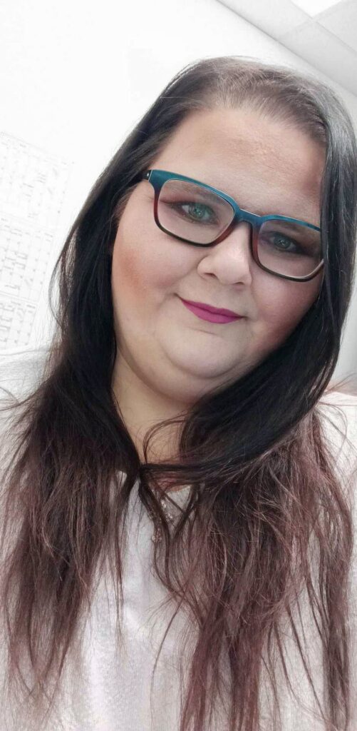 The photo shows a person with long dark hair wearing teal-framed glasses and a light-colored top. They are indoors, in and office type setting. The background includes a white wall, a ceiling with fluorescent lights. The person is facing the camera and appears to be taking a selfie.