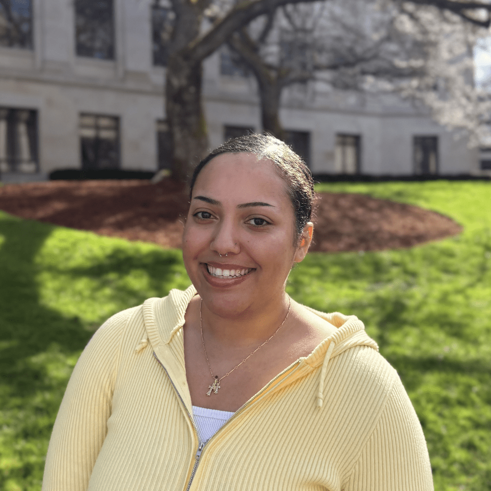 Person smiling at the camera outside on a sunny day. They are wearing a light yellow jacket and there is grass, trees, and a building behind them.