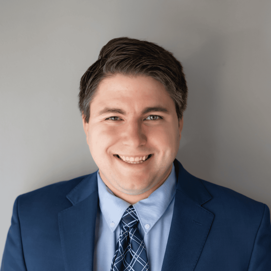 Professional headshot of Cody Clark smiling at the camera, wearing a navy suit, light blue dress shirt, and patterned tie against a neutral gray background.
