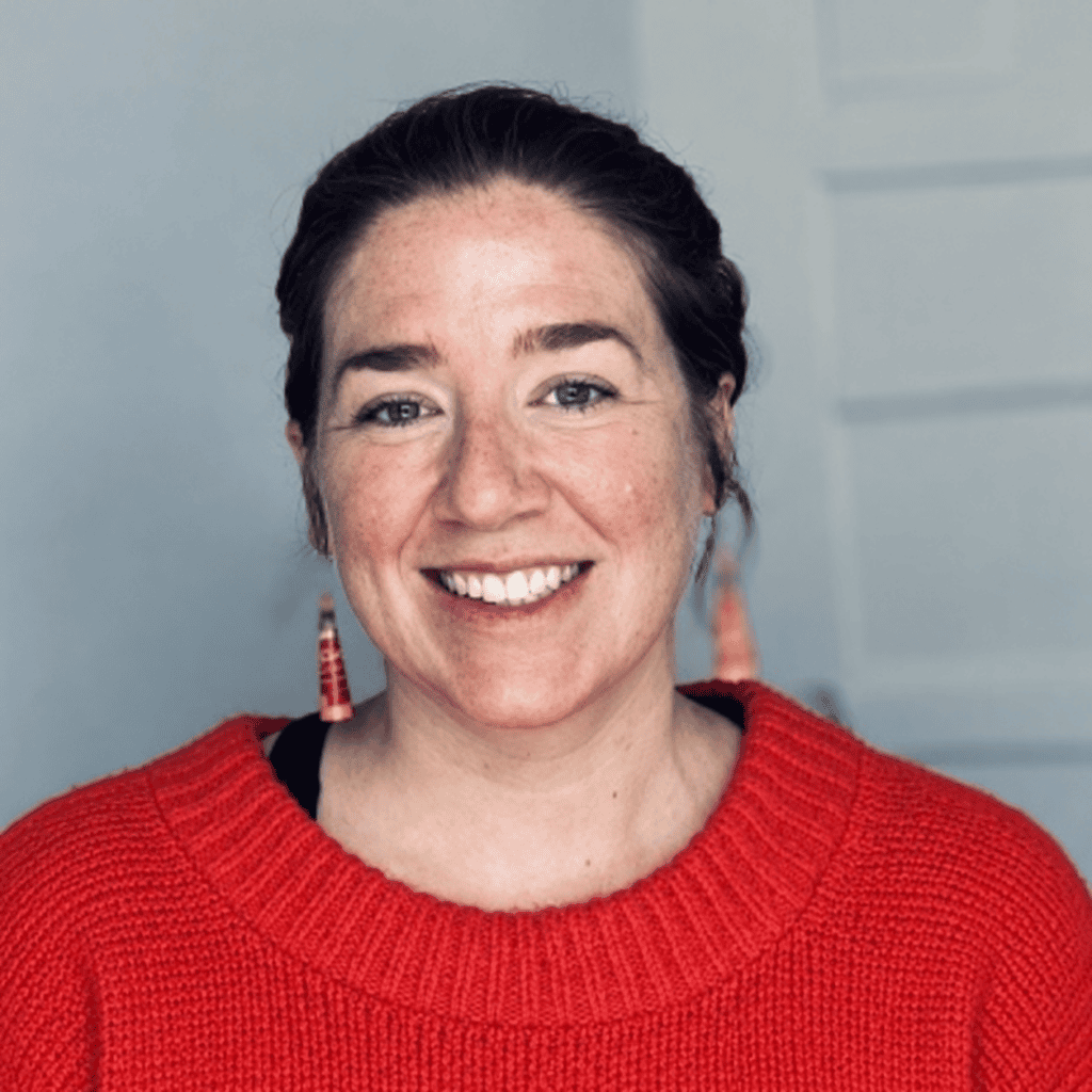 Headshot of Amanda smiling, with dark hair pulled back, colorful dangling earrings, and a red sweater, against a grey background.